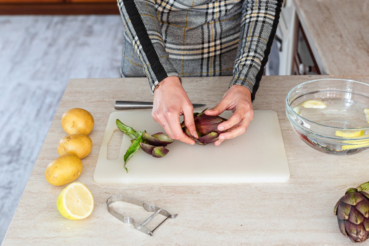 Mani di donna che puliscono carciofi su tagliere di plastica con vicino patate ancora da tagliare
