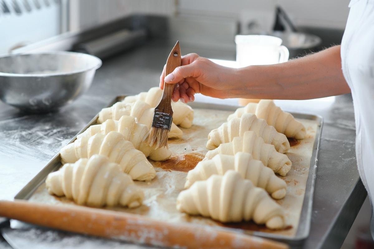 Cornetti o croissant lievitati su teglia da forno vengono spennellati con latte