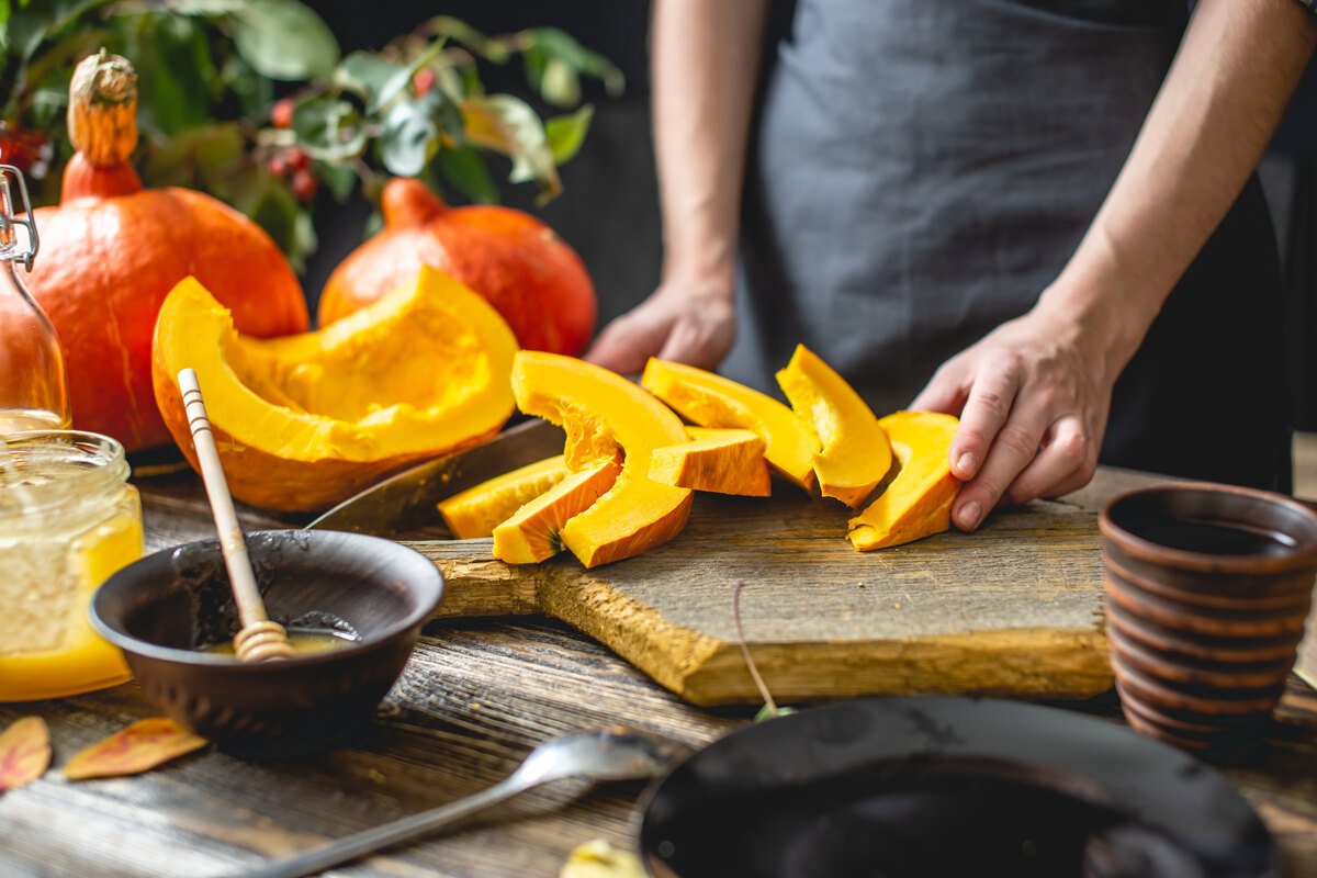Fette di zucca su tagliere di legno con miele in primo piano e donna con grembiule sullo sfondo.
