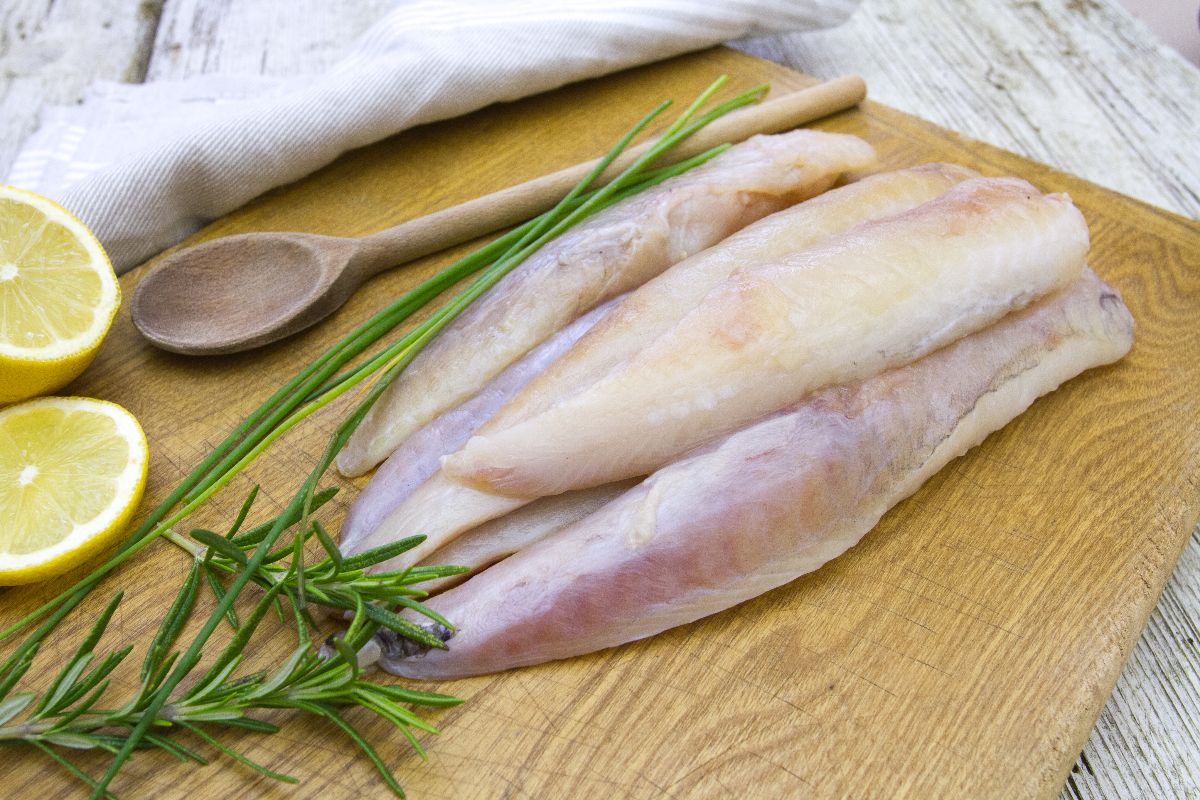 Filetti di rana pescatrice su tagliere di legno con vicino cucchiaio, erbe aromatiche e limone.