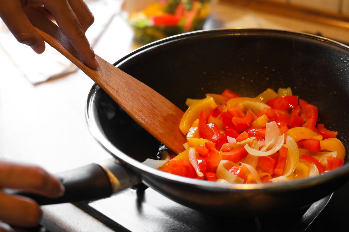 Padella con peperoni e cipolle in cottura mescolati con cucchiaio di legno