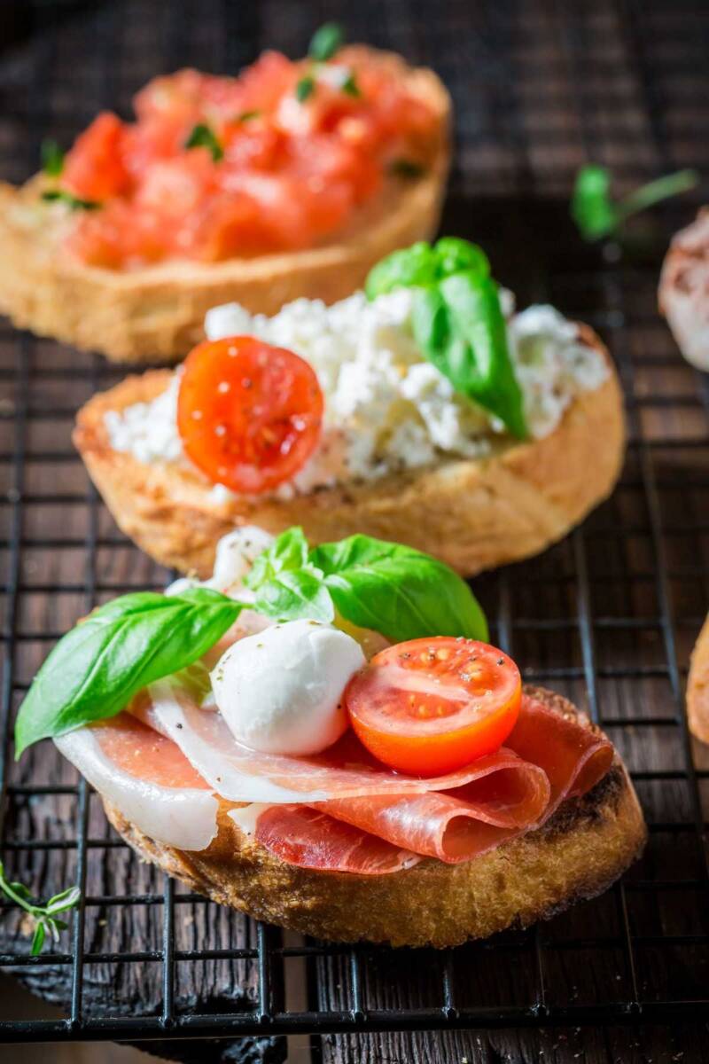 Tris di bruschette con in primo piano crudo, mozzarella e pomodoro, dietro ricotta e pomodoro, sullo sfondo con pomodorini e basilico