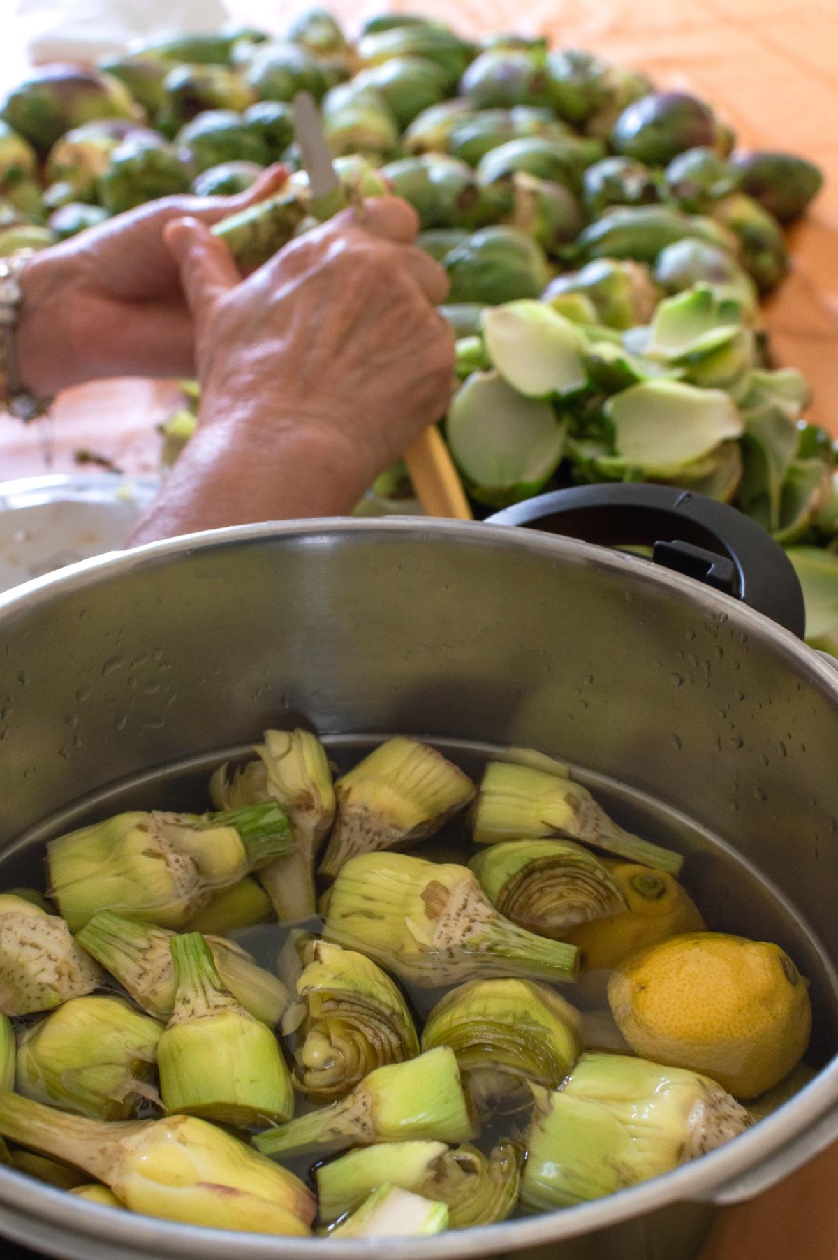 Carciofi puliti in pentola acidulata con succo di limone e mani sullo sfondo che puliscono altri carciofi