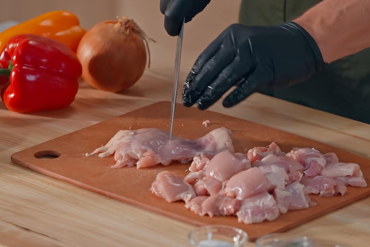 Mani con guanti neri che tagliato a tocchetti carne di pollo con peperoni sullo sfondo