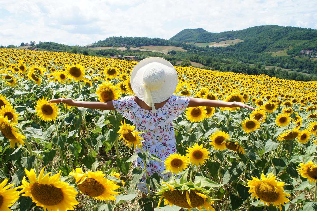 Ragazza di spalle con vestito a fiori e cappello di paglia che cammina tra girasoli.