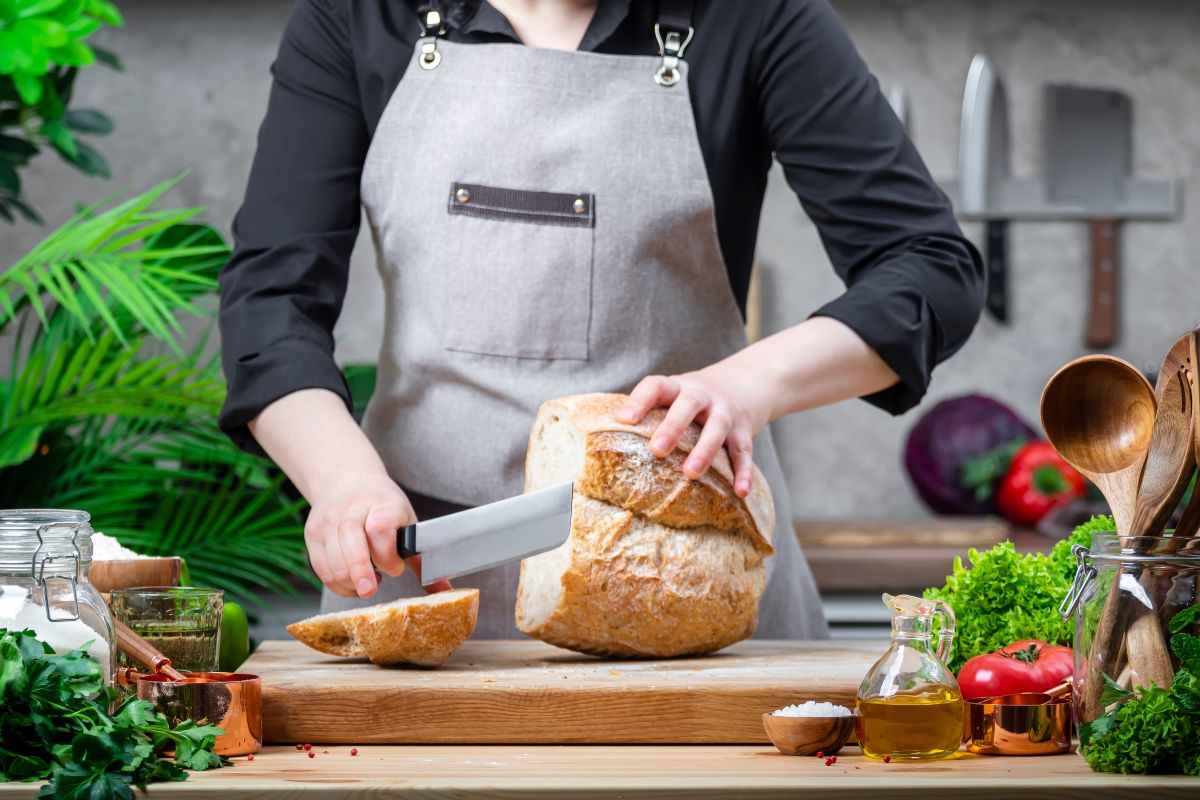 La chef in cucina intenta a tagliare il pane tra ingredienti freschi.