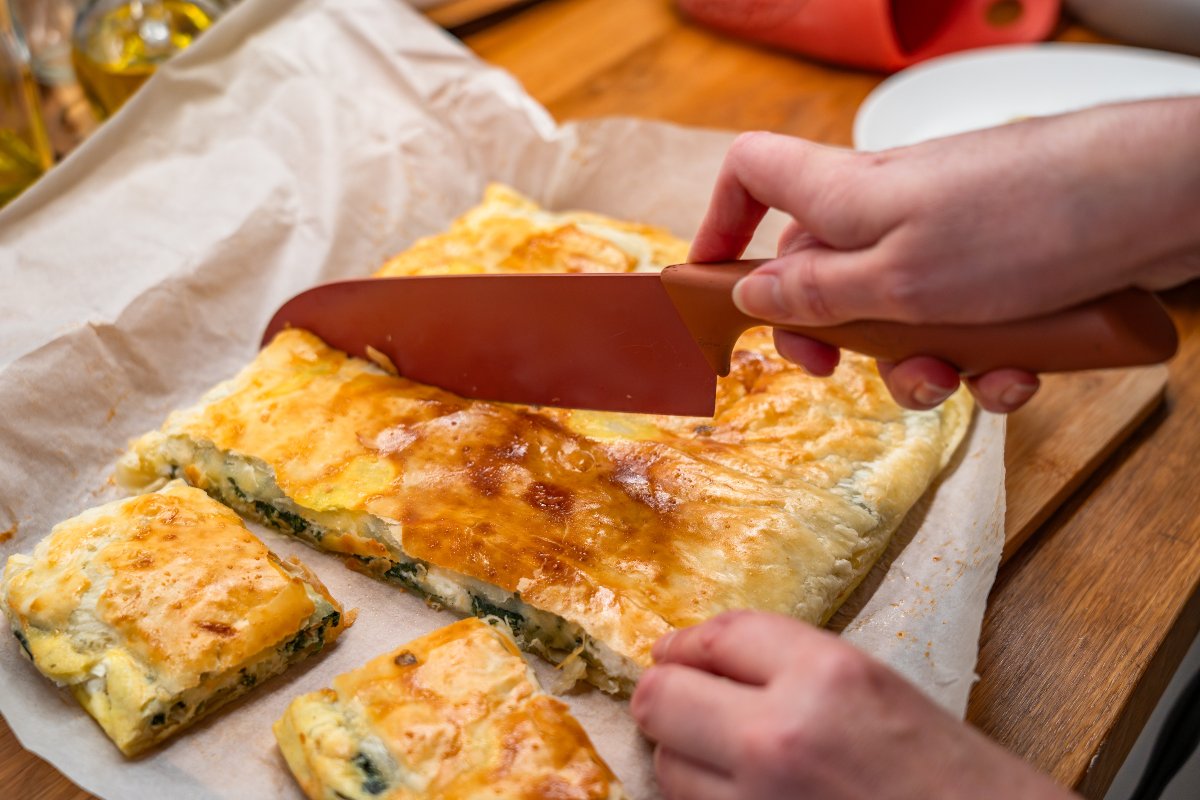 Una torta salata con spinaci e feta sulla sua carta forno mentre viene tagliata in quadretti.