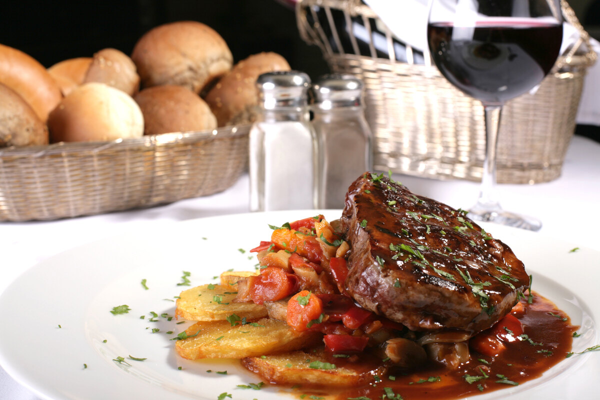 Piatto di braciole cotte nel vino con patate e carote. Sullo sfondo pane e calice di vino rosso.
