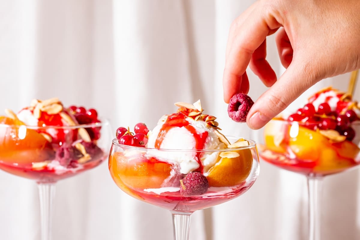 Mano che aggiunge lampone a coppetta con gelato, pesche sciroppate e coulis di lamponi