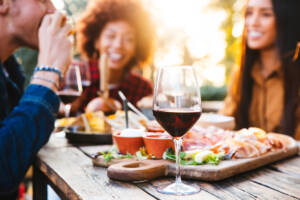 Primo piano con bicchiere di vino, tagliere di salumi e sullo sfondo donne che ridono