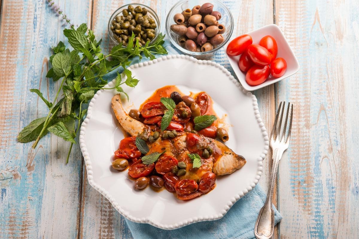 Piatto con trancio di pesce spada con olive, pomodorini e capperi e vicino ingredienti utilizzati