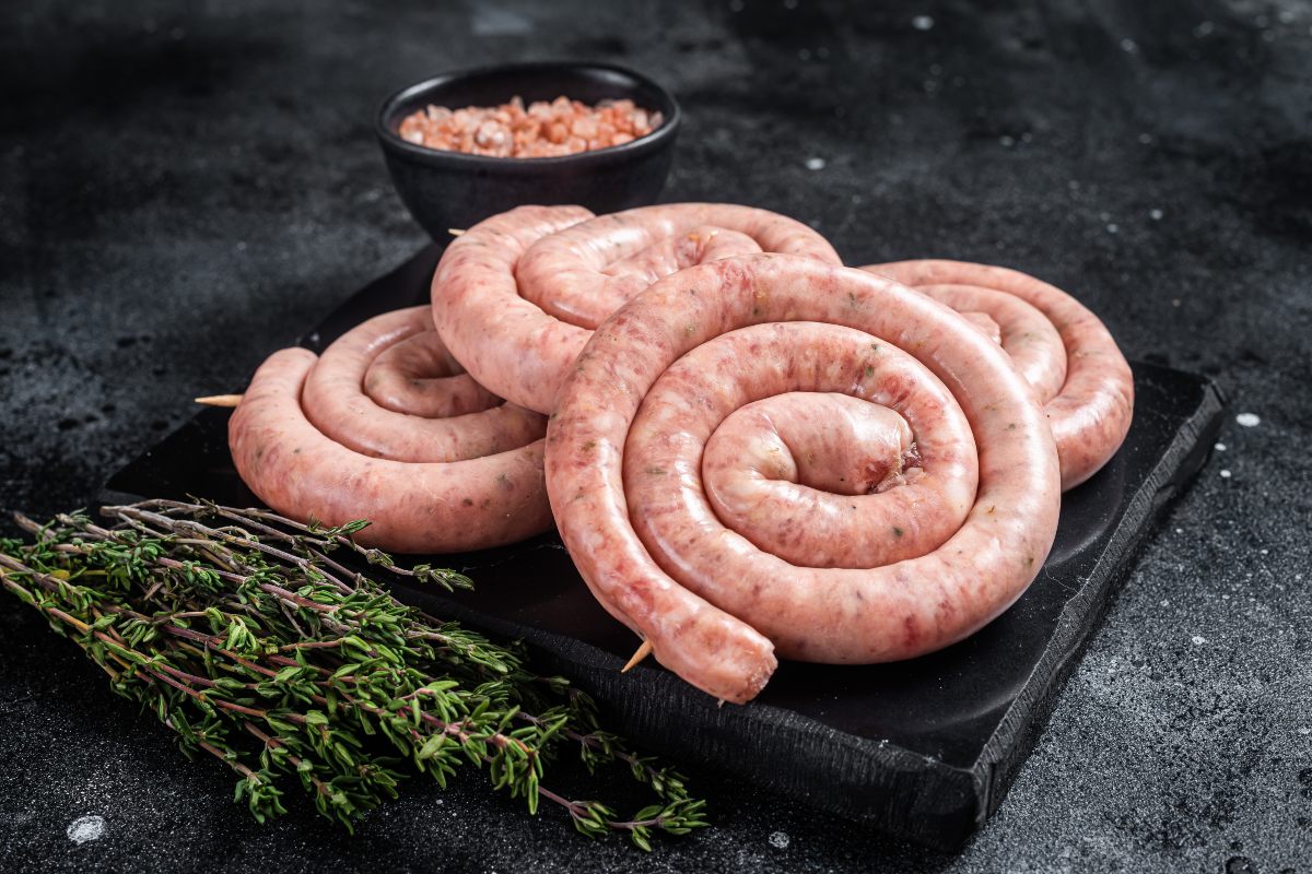 Raw Luganiga sausage rolled and held with skewers on a slate cutting board accompanied by aromatic herbs.