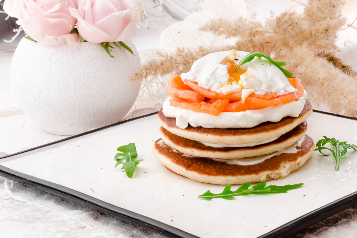 Pila di pancake salati con strati di formaggio e salmone su piatto piano.