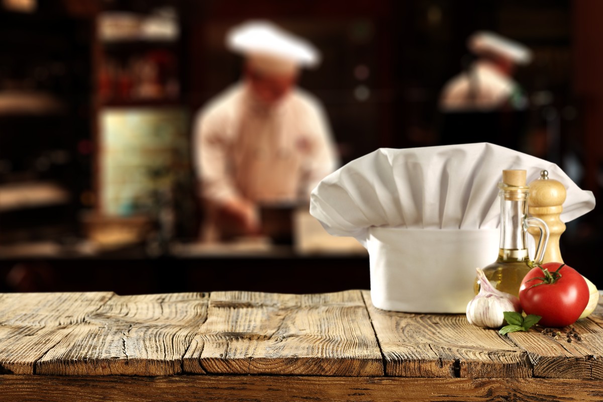 Un cappello da chef, una bottiglia d'olio, un pomodoro, del sale e dell'aglio si un piano di legno dentro un ristorante