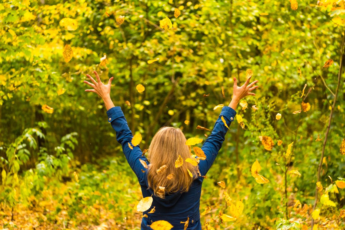 Donna di spalle con le braccia alzate, immersa in un bosco autunnale.